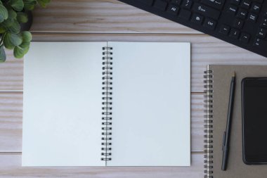 Siyah bilgisayar klavyesi, kalem, not defteri, akıllı telefon ve saksı bitkisi olan ofis masasının tam üstünde.