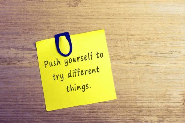 İlham verici alıntılar - Tahta masanın tepesinde mavi ataç ve sarı çıkartma üzerinde farklı şeyler denemek için kendinizi zorlayın.