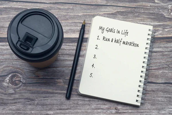 My goals in life list on notepad with pen and disposable coffee cup on wooden desk.