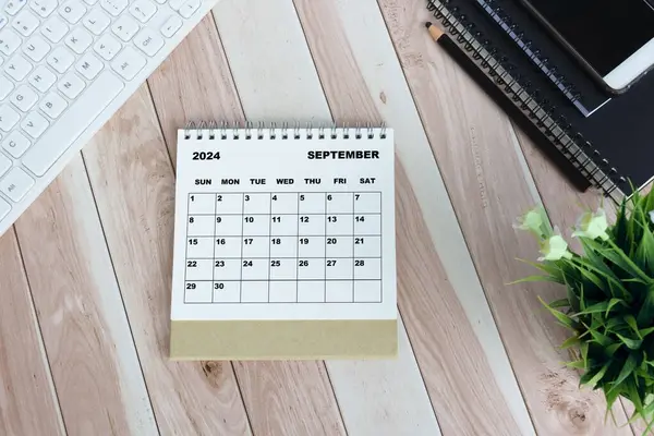 White September 2024 calendar on wooden desk with keyboard, note books, pencil, potted plant and smartphone.
