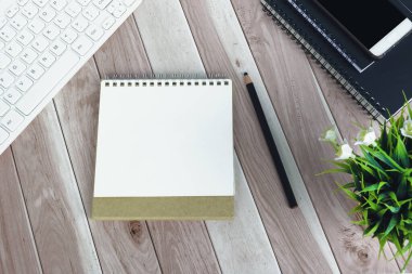 Beyaz bilgisayar klavyesi, kalem, akıllı telefon, not defteri ve saksı bitkisi olan ofis masasının tam üstünde.