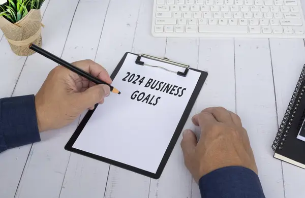 Hands writing a 2024 business goals on a clip board on white wooden desk. Business and finance concept.
