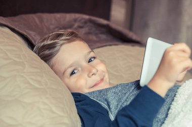Smiling little boy with touchpad lying in bed and looking at camera. 