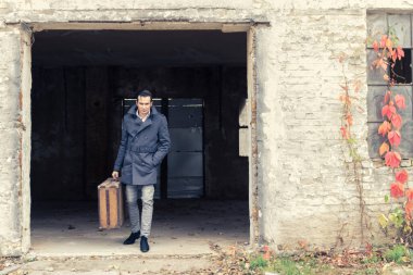 Determined man carrying suitcase while exiting ruined building and looking at camera. 