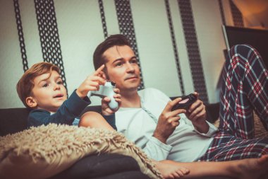 Small kid and his dad playing video games while relaxing on the sofa in the living room.