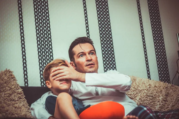 Shocked father covering son\'s eyes while watching something inappropriate on TV at home.