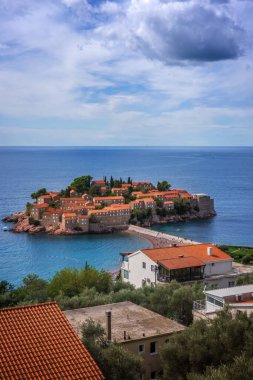Karadağ 'ın Budva kenti yakınlarındaki güzel Sveti Stefan adası. Mavi Adriyatik Denizi 'ndeki adanın tarihi kırmızı çatılı evleri, parçalı bulutlu bir gökyüzünün altında dar bir isthmus ile kıyıya bağlıdır.