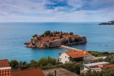 Karadağ 'ın Budva kenti yakınlarındaki güzel Sveti Stefan adası. Mavi Adriyatik Denizi 'ndeki adanın tarihi kırmızı çatılı evleri, parçalı bulutlu bir gökyüzünün altında dar bir isthmus ile kıyıya bağlıdır.