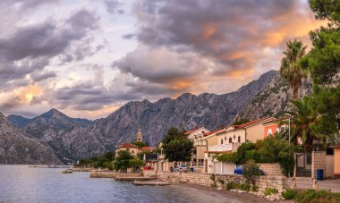 Karadağ 'ın Kotor kentindeki Dobrota' nın sabah manzarası. Fotoğraf, kıyı boyunca sakin suları, yemyeşil binaları ve bulutlu, gün doğumunda gökyüzünün altında engebeli dağları yakalar.