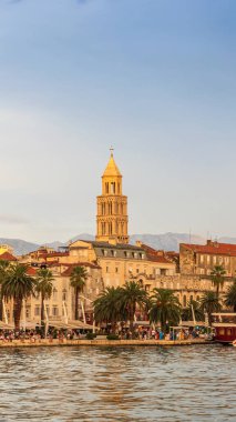 Hırvatistan 'ın Split kentindeki canlı Riva gezinti alanında gün batımı. Palmiye ağaçları ve tarihi binalarla dolu rıhtım insanlarla ve renkli teknelerle dolu..