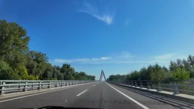 Açık gökyüzünün altında, yeşil ağaçlarla çevrili bir köprüye yaklaşan bir araba, Hırvat yolu boyunca ilerliyor. Sahne, modern köprünün manzaralı doğasıyla çevrili pürüzsüz sürücüsünü yakalıyor..