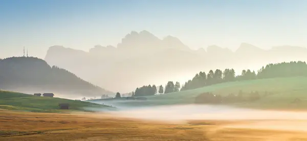This photo captures Alpe di Siusi in the Dolomites at sunrise, with mist rolling over green meadows, scattered cabins, and distant mountains bathed in soft morning light