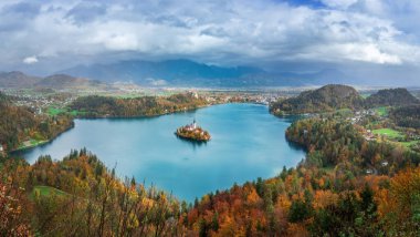 Bled Gölü 'nün bu çarpıcı manzarası turkuaz sularını, pitoresk bir adayı, tarihi bir kiliseyi, bereketli ormanları, yuvarlanan tepeleri ve dramatik bir gökyüzünün altındaki sisli dağları yakalar..