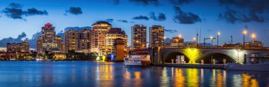 Batı Palm Beach, Florida kıyısı gün batımından sonra aydınlık ufuk çizgisi, marina, yat ve sakin suya yansıyan yansımalar, kentsel ışıklar, modern binalar ve dramatik akşam gökyüzü..