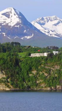 Yeşil yemyeşil tepeleri ve karla kaplı dağları olan Norveç kıyı şeridinin manzarası fiyorttan geçen bir yolcu gemisinden çekiliyor. 