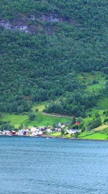 Yeşil tepeleri ve dağları yemyeşil olan Norveç kıyı köyünün manzarası fiyorttan geçen bir yolcu gemisinden çekiliyor. 