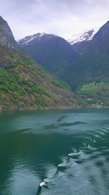 Norveç fiyortlarının sakin sularında seyreden bir geminin arkasında dalgalar yumuşak güneş ışığı altında yüzeyde ritmik ve yansıtıcı bir desen oluşturuyor..
