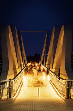 Ukrayna 'nın başkenti Kyiv' deki Azov yaya köprüsünün gece görüşü. Derin mavi gökyüzüne karşı sıcak ışıklar altında hareket eden insan siluetleri..