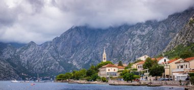 Geleneksel Akdeniz evleri, kilise kulesi ve güneşli bir günde sakin Kotor Körfezi 'nin üzerinde yükselen dağlarla Karadağ' ın Dobrota kentinin manzarası.
