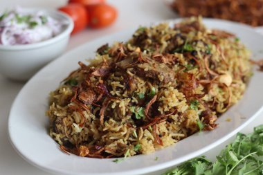Baharatlı yahni pilavı ile hazırlanmış koyun eti biryani yanında bol miktarda karamelli soğan ve yoğurt ile marine edilmiş koyun eti. Yoğurt soğan salatasıyla servis edilir.