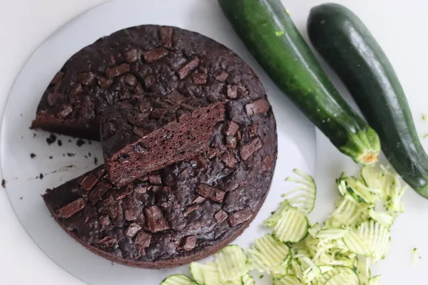 Slice of Zucchini chocolate cake on top of the round cake. Moist double chocolate cake with grated zucchini, coco powder, chocolate and chocolate chips. Shot on white background along with zucchinis
