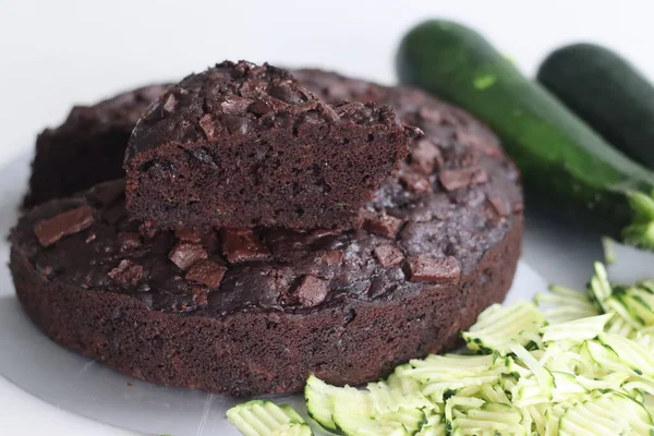 Slice of Zucchini chocolate cake on top of the round cake. Moist double chocolate cake with grated zucchini, coco powder, chocolate and chocolate chips. Shot on white background along with zucchinis