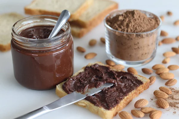 Bread slice with almond chocolate spread, a heavenly blend of rich almonds and smooth chocolate. Shot with many almonds, a bottle of almond chocolate spread and a bowl of coco powder in the background