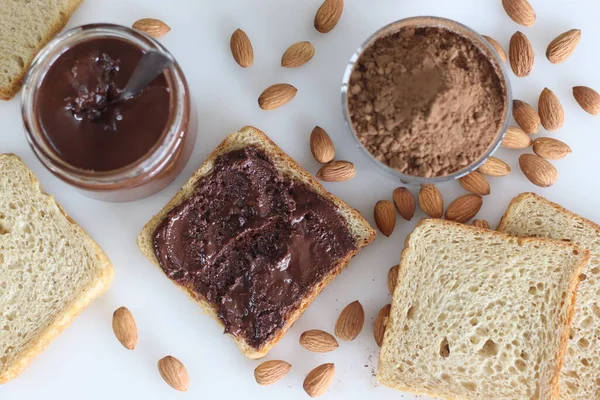 Bread slice with almond chocolate spread, a heavenly blend of rich almonds and smooth chocolate. Shot with many almonds, a bottle of almond chocolate spread and a bowl of coco powder in the background