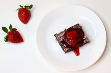 Üstü taze çilek ve nane yapraklarıyla kaplı çikolatalı brownie, beyaz üzerine zarif bir tabaka. Bir kase çilek sosu ve arka planda tam çilek, tatlı ve yemek fotoğrafçılığı için ideal.