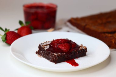 Üstü taze çilek ve nane yapraklarıyla kaplı çikolatalı brownie, beyaz üzerine zarif bir tabaka. Bir kase çilek sosu ve arka planda tam çilek, tatlı ve yemek fotoğrafçılığı için ideal.