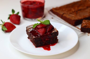 Üstü taze çilek ve nane yapraklarıyla kaplı çikolatalı brownie, beyaz üzerine zarif bir tabaka. Bir kase çilek sosu ve arka planda tam çilek, tatlı ve yemek fotoğrafçılığı için ideal.