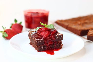 Üstü taze çilek ve nane yapraklarıyla kaplı çikolatalı brownie, beyaz üzerine zarif bir tabaka. Bir kase çilek sosu ve arka planda tam çilek, tatlı ve yemek fotoğrafçılığı için ideal.
