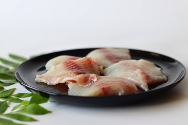 Kırmızı damarlı taze tilapia filetoları, taze köri yapraklarıyla çevrili yuvarlak siyah seramik bir tabakta ustaca düzenlenmiş. Asya yemekleri, deniz ürünleri tarifleri ve sağlıklı yemek hazırlamak için mükemmel.