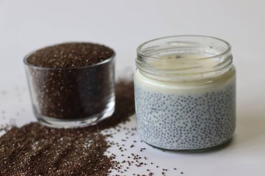 Kavanozda Chia 'ya batırılmış süt ya da chia pudingi, etrafa saçılmış chia tohumları ve bir kap kuru tohumla çevrili. Sağlıklı görseller, kahvaltı temaları ve beslenme içeriği için iyi bir kremalı vegan yemeği.