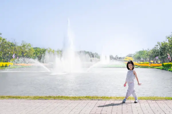 Asian Girl Strolling by Park Fountain on Sunny Day, Kid in dotted outfit takes leisurely stroll by majestic fountain in the park, with sun casting a soft glow on serene landscape. Child aged 6 years.