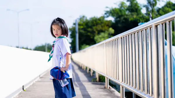 A schoolgirl walks home from school in the hot summer sun, a girl frowns against the hot sun, Child walks on an overpass, looks back at the camera, a 6 year old Kid wears a Thai school uniform.