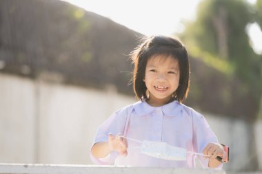 Elinde tıbbi maskeyle parkta neşeyle oynayan gülümseyen bir çocuğun portresi, tatlı gülümseyen kız, beyaz okul üniforması giyen liseli kız, yaz veya bahar akşamları, keyifli bir ruh hali..