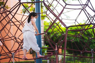 Kız oyun parkında ip oyun ekipmanına tırmanırken eğleniyor. Çocuk sağlığını korumak için maske takıyor. Oyun ekipmanları, çocukların tırmanması için üç boyutlu bir şekilde örülmüş bir halattır..