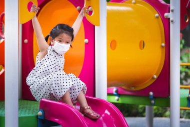 Çocuklar Oyun Sahasında Mutlulukla Oynuyor. Asyalı güzel kız kaydırağın tadını çıkarıyor. Sevimli küçük çocuk sallan ve neşeyle takıl. Virüsü ve toksik tozu korumak için tıbbi maske takan çocuk.