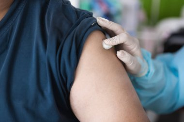 Hastane ortamında bir hastayla ilgilenen bir doktor. Bir elinde şırınga, üst kolunda aşı olan birinin yakın çekimi, sağlık ve kişisel bakım elementleriyle çevrili..