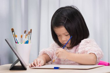 Küçük bir kız renkli sanat malzemeleriyle çevrili bir masada boya fırçasıyla mutlu bir şekilde resim çiziyor. 3-4 yaşlarında Asyalı bir çocuk. Evde eğitim. Çocuk resim öğreniyor.