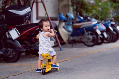 Çocuk arka arkaya park etmiş motosikletlerle dolu bir yolda sarı bir scooter sürüyor yaz ya da bahar zamanı tatlı tatlı gülümseyen bir çocuk. Yolda oynarken dikkat ve güvenlik kavramı.