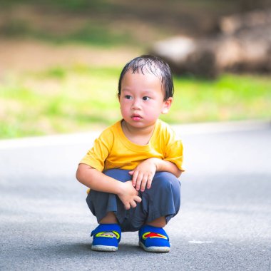 Asyalı çocuğun kafası sıcak ve nemli havadan dolayı terden sırılsıklam olmuş. Çocuk yolun ortasında yürürken dinlenmek için çömeliyor. 2 yaşındaki çocuk koyu sarı bir gömlek giyiyor..