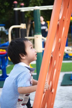 Küçük çocuk yaz güneşinde çocuk parkında mutlu bir şekilde oynuyor. Asyalı çocuk, küçük ve büyük kasların tırmanma, oynama ve geliştirme, karar verme ve cesaret için turuncu oyun ekipmanlarına tırmanıyor..