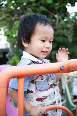 Güneşli bir günde çocuk parka tırmanırken oyuncakla oynarken gülümseyen küçük çocuk. Çocuk baharı ya da yazı dışarıda geçirmeyi ve tırmanmayı seviyor. 2 yaşında Asyalı bir çocuk. Dikey resim.