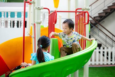 Çocuklar güneşli bir günde oyun alanında birlikte oynarken eğleniyorlar. Kız ve erkek kardeş mutlu bir şekilde kaydırağın üzerinde oynuyorlar. Aile zamanı..