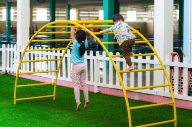 Çocuklar oyun alanında oynuyorlar, sarı kemerli köprü merdivenlerine tırmanıyorlar..