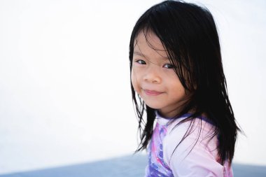 Uzun, ıslak siyah saçlı, renkli mayo giyen neşeli Asyalı bir çocuğun parlak, yüksek anahtarlı portresi. Kız kurnazca gülümsüyor. Temiz beyaz arka plan, yaz eğlencesi ve çocukluk masumiyeti..