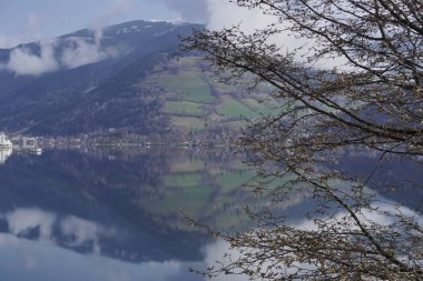 Avusturya Alpleri 'nde yer alan Zell am See' nin bulutlu hava günlerinde bankları kapatın. Arkaplanda dağlar ve şehir, saf suyun üzerinde yansıyor.