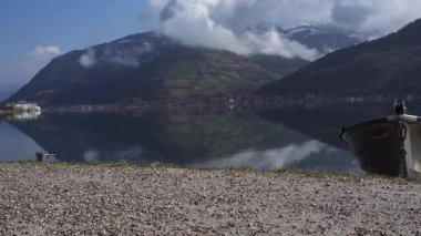 Zell am See, Avusturya 'da Zell Gölü yakınlarında siyah pantolonlu ve kirli ayakkabılı bir turist yürüyor. Metal bir tekne kristal berrak suda yüzüyor, çarpıcı alp manzarasını yansıtıyor.